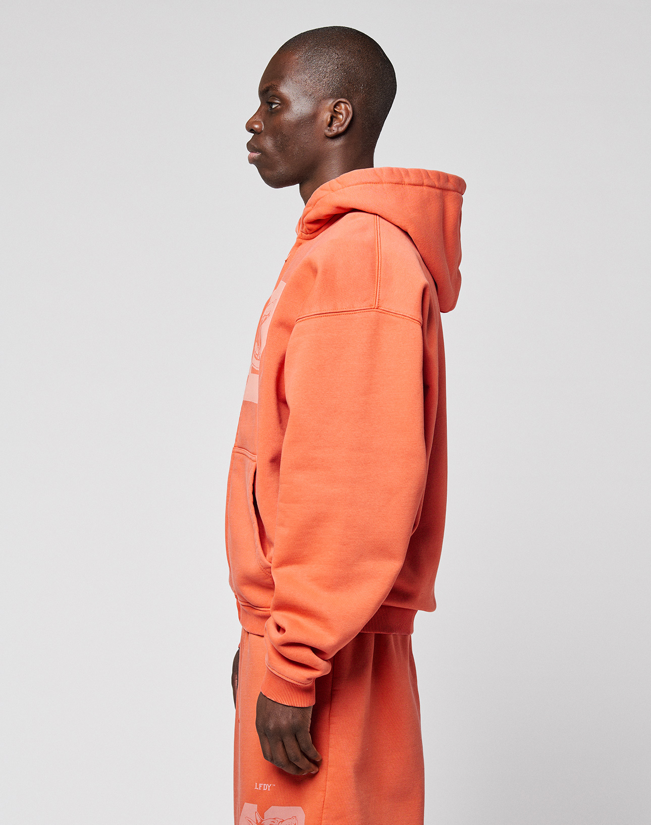 A person stands in profile against a plain background, wearing an orange LFDY 40 Hooded Zip by LIVE FAST DIE YOUNG with matching sweatpants.