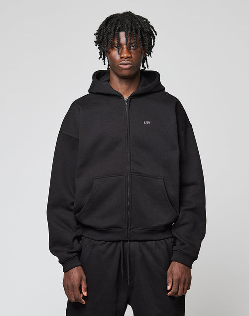 A person with medium-dark skin and short, twisted hair wears the LIVE FAST DIE YOUNG Basic Hooded Zip in black with matching pants, standing against a plain light gray background.