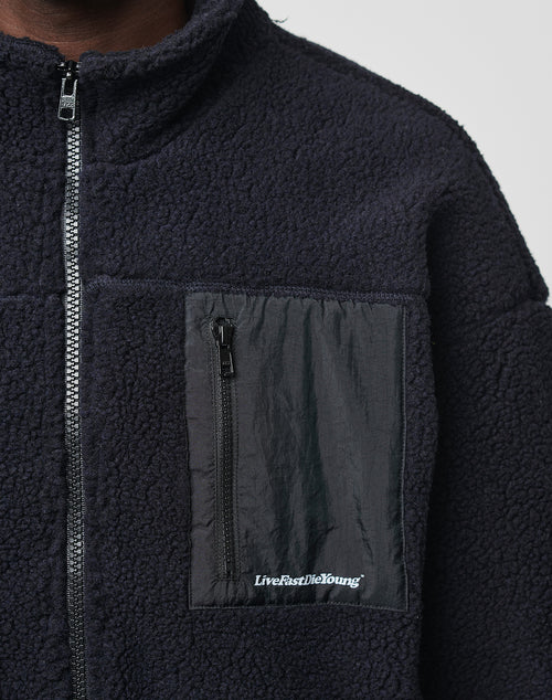 Close-up of the Basic Teddy Jacket by LIVE FAST DIE YOUNG: black fleece, stand-up collar, zip front, regular fit, and a nylon chest pocket with “Live Fast Die Young” printed in white at the bottom.