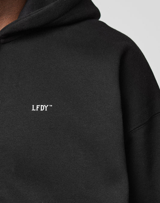 Close-up of a person wearing the LIVE FAST DIE YOUNG Basic Hooded Zip in black, featuring a white “LFDY™” logo on the chest. The image highlights the branding and shows part of the neck in a slightly cropped fit.