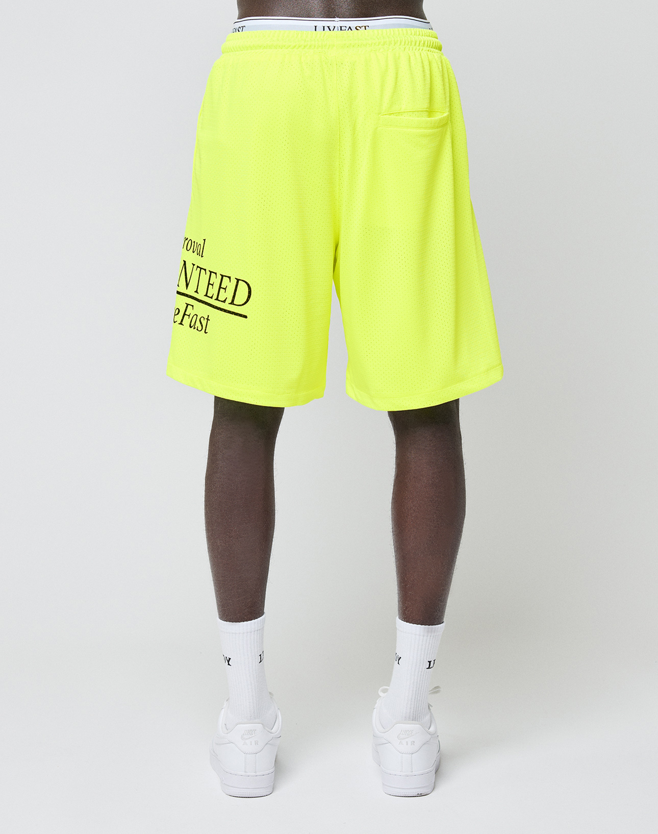 A person is shown from the back against a white background wearing LIVE FAST DIE YOUNG's LFDY Guaranteed Mesh Shorts in bright yellow (wide fit, elastic waistband), white socks, and white sneakers. Partially visible black text appears on the shorts' left leg.
