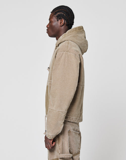 A person with braided hair stands in profile against a plain background, wearing a tan Faded Workwear Jacket by LIVE FAST DIE YOUNG with a hood and matching pants featuring visible seams, pocket details, and a practical kangaroo pocket.