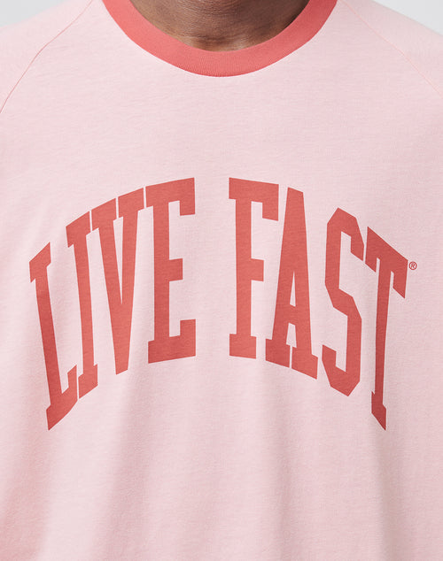 Close-up of a person in the LIVE FAST DIE YOUNG Live Fast Contrast Tee, a soft light pink shirt featuring LIVE FAST printed boldly in red capital letters across the chest.