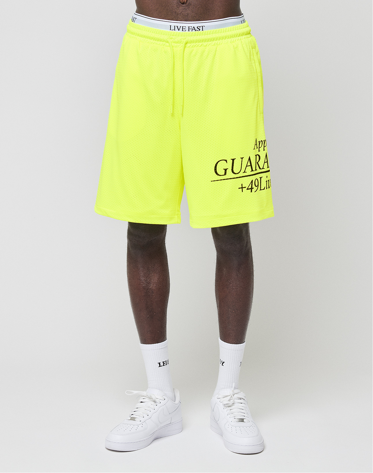 A person wears bright yellow LFDY Guaranteed Mesh Shorts by LIVE FAST DIE YOUNG, featuring a wide elastic waistband with "LIVE FAST" text, paired with white socks and sneakers, standing against a plain white background.