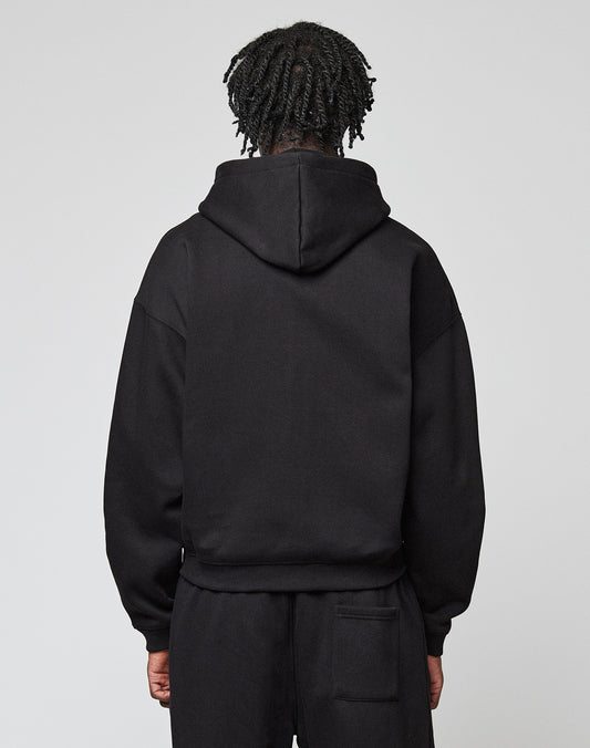 A person with short, twisted hair faces away from the camera, wearing a black LIVE FAST DIE YOUNG Basic Hooded Zip sweatshirt and black pants against a plain light background.