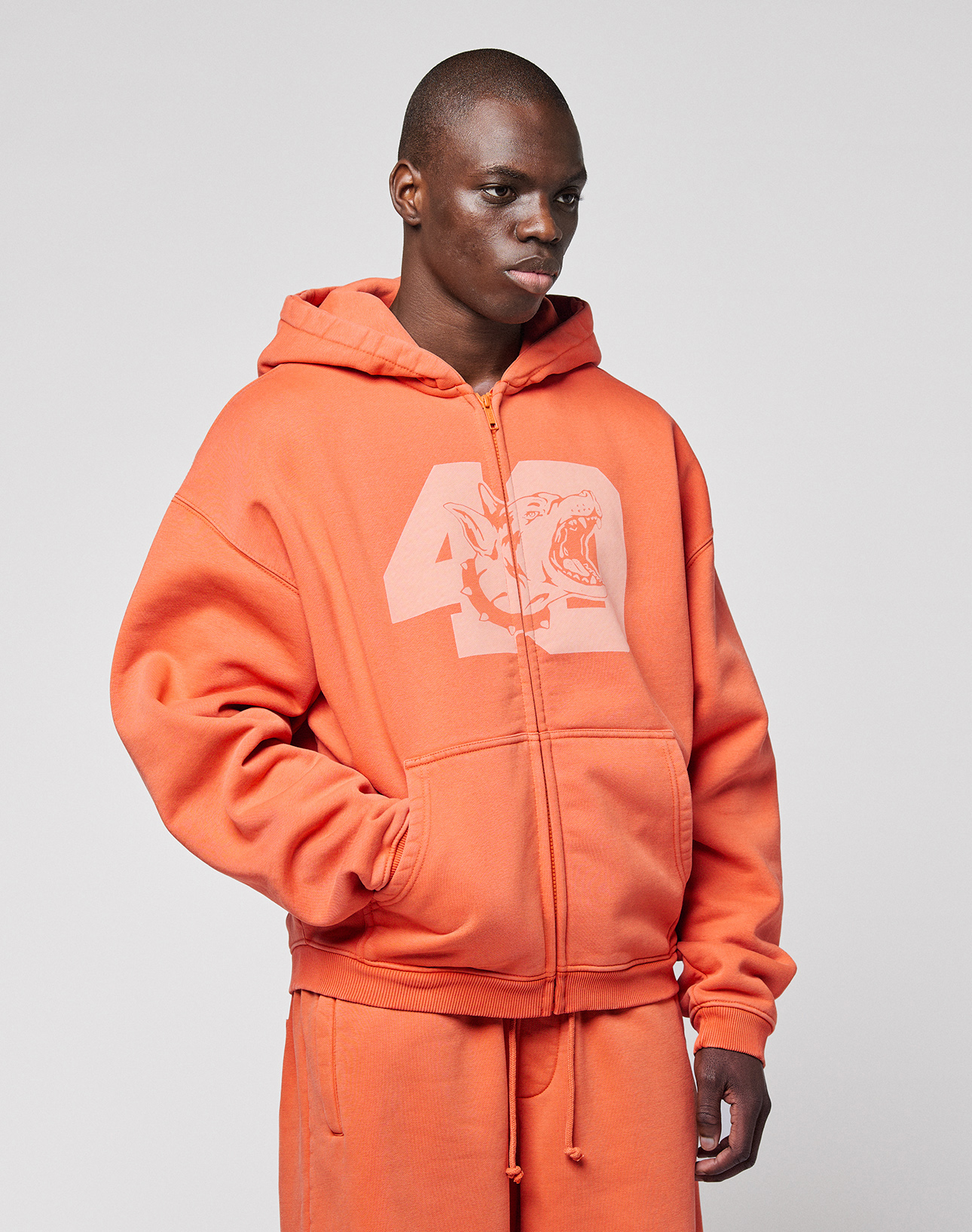 Someone wearing the LFDY 40 Hooded Zip by LIVE FAST DIE YOUNG, an oversized orange zip-up pullover with a frontprint graphic number 42 and animal head, paired with matching orange sweatpants, stands against a plain light gray background.