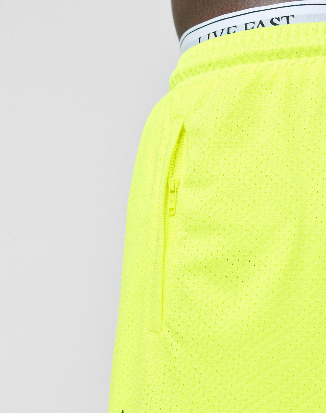 Close-up of a person wearing LFDY Guaranteed Mesh Shorts by LIVE FAST DIE YOUNG in neon yellow. These wide fit polyester shorts feature a zippered side pocket and an elastic waistband. Branded LIVE FAST underwear is visible; the background is plain and light.