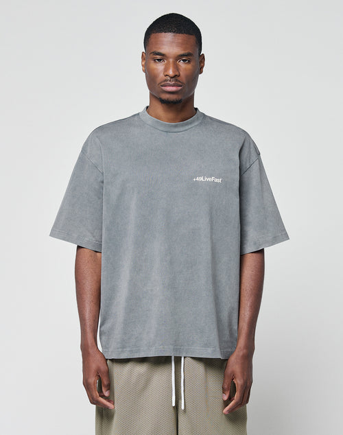 A man stands against a light gray background, wearing the LIVE FAST DIE YOUNG Basic +49 Chest Tee—an oversize, faded gray 100% Baumwolle shirt with a small chest logo—paired with light brown mesh shorts. He faces the camera neutrally.