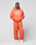 A person stands against a plain backdrop wearing an orange LIVE FAST DIE YOUNG Oversize Fit zip-up hoodie and LFDY 40 Wide Cuff Jogger with Druck auf dem Bein, seitliche Eingrifftaschen, and white sneakers. Hands in pockets, expression neutral.