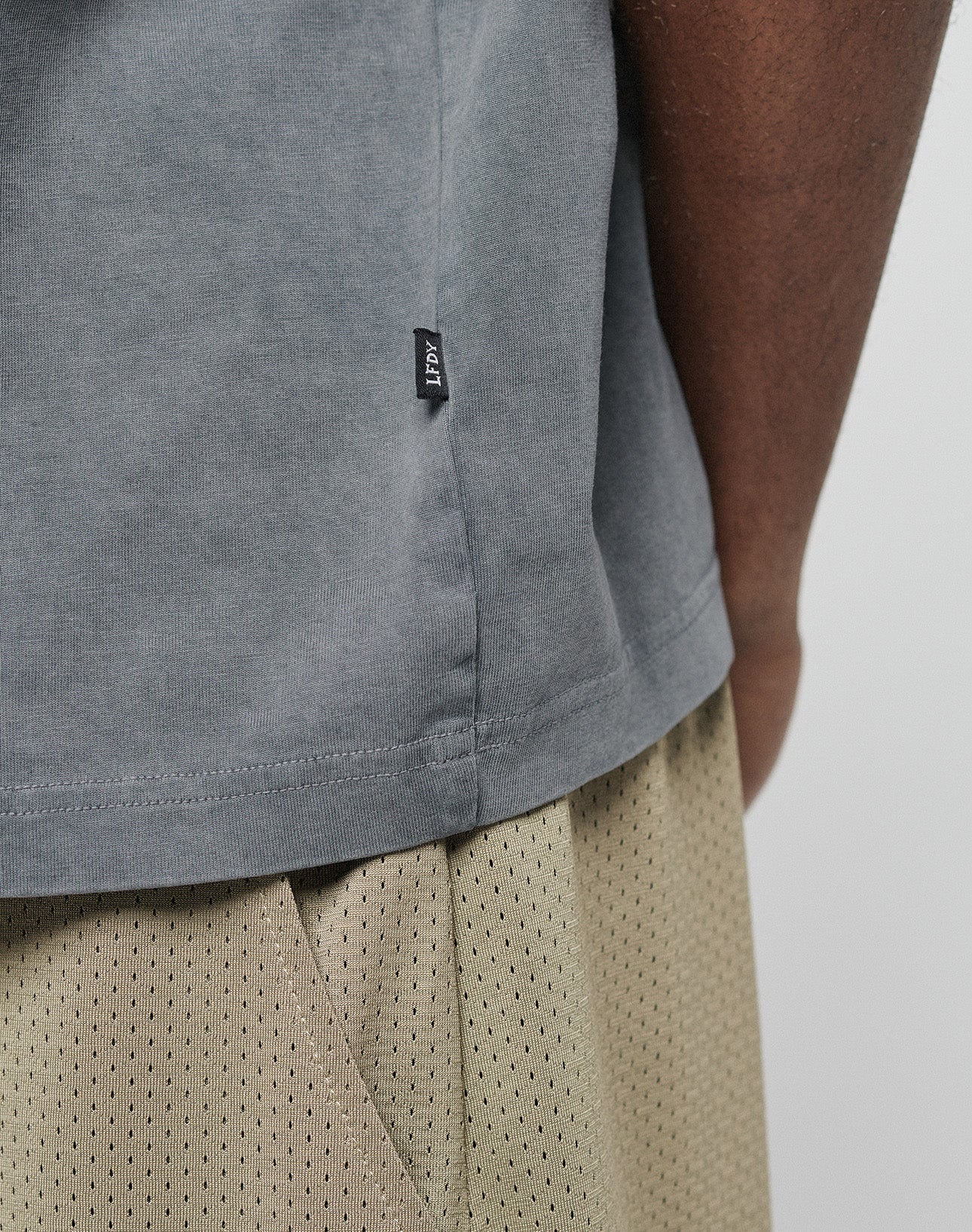 Close-up of a person in the LIVE FAST DIE YOUNG Basic +49 Chest Tee in grey (100% Baumwolle) with a small black seam tag, subtle logo print, and beige mesh shorts. The oversize fit tee is shown against a plain background.