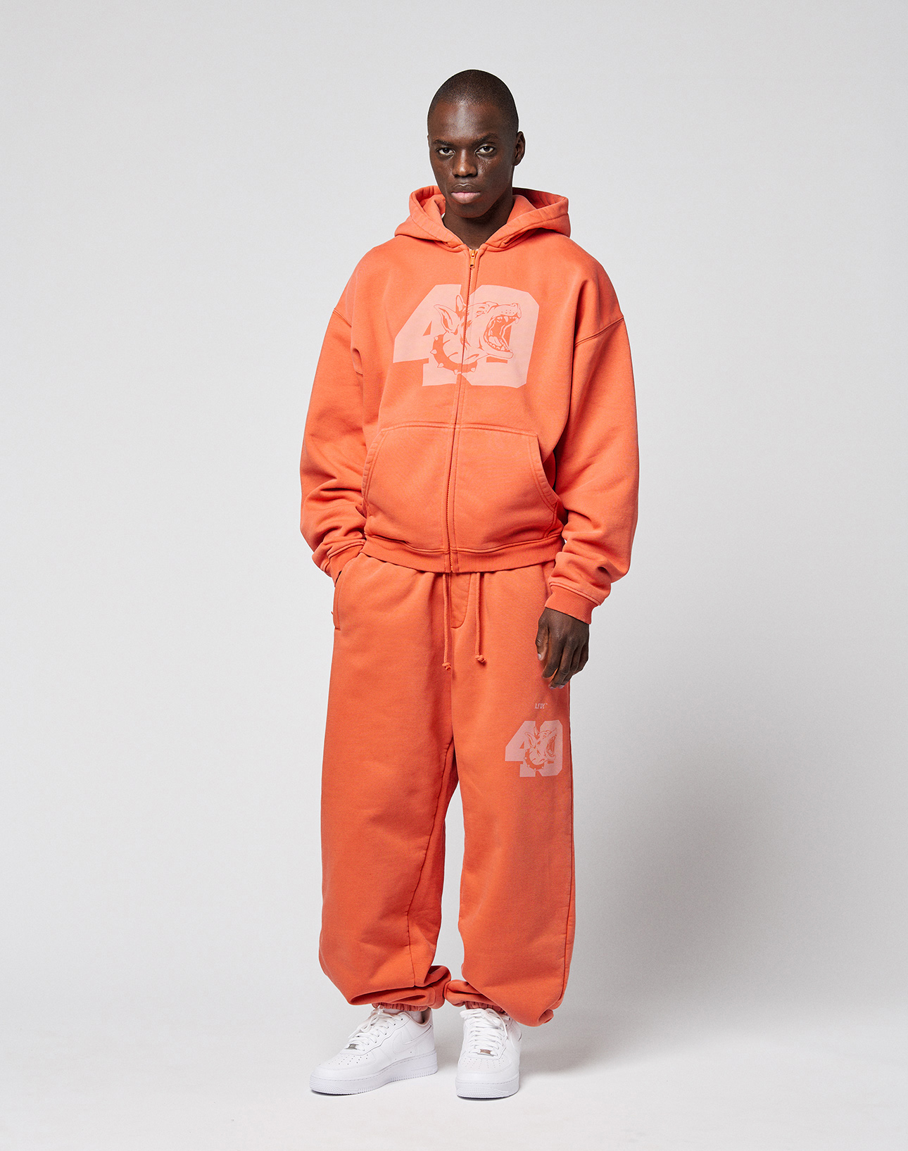 A person stands against a plain background wearing the orange LFDY 40 Hooded Zip by LIVE FAST DIE YOUNG with matching sweatpants featuring white frontprint graphics and white sneakers. Hands in pockets, they look at the camera.
