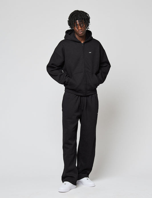 A person with short, curly hair wears the LIVE FAST DIE YOUNG Basic Hooded Zip and matching black sweatpants, standing hands-in-pockets against a plain white background with white sneakers.