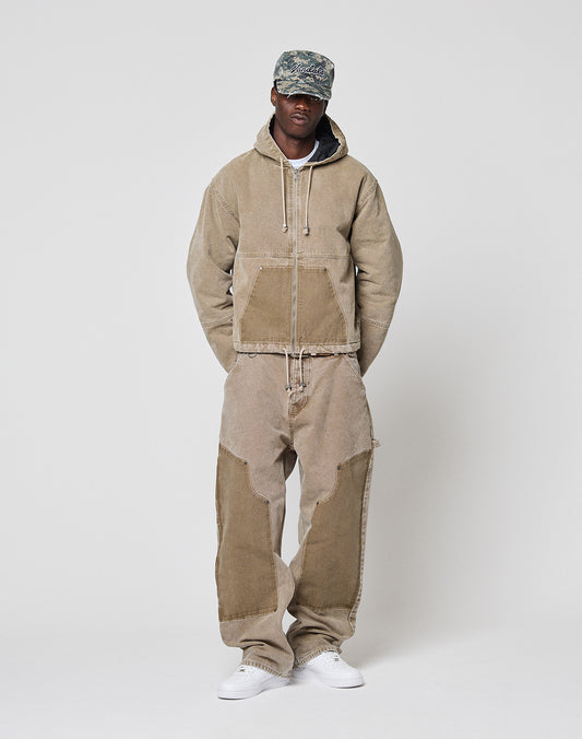 A person in a tan Faded Workwear Jacket by LIVE FAST DIE YOUNG, loose corduroy pants with contrasting knee patches, a camouflage cap, and white sneakers stands against a plain light background.