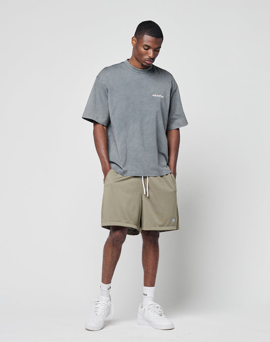 A man stands against a plain background wearing the LIVE FAST DIE YOUNG Basic +49 Chest Tee in grey (oversize fit, 100% Baumwolle), khaki shorts with white drawstrings, white socks, and white sneakers. His head is slightly tilted down with hands in his pockets.