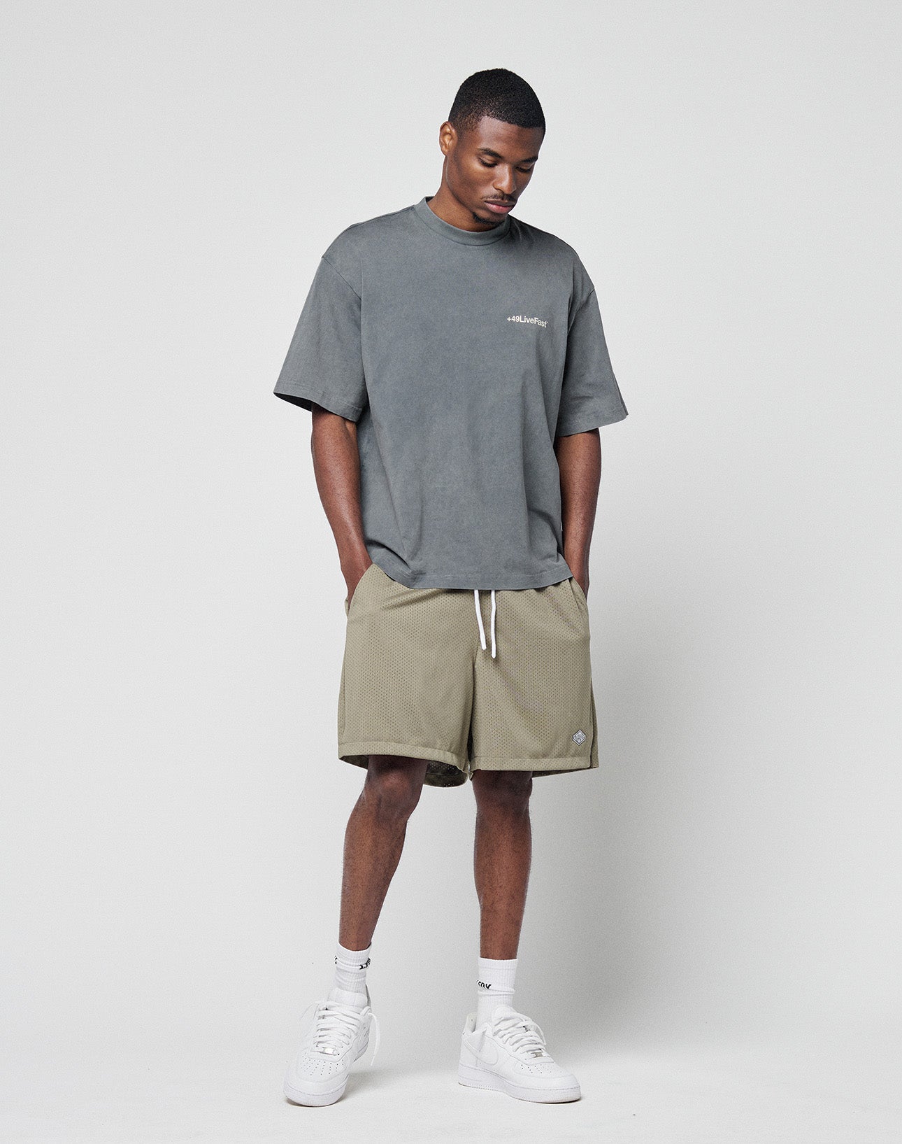 A man stands against a plain background, wearing the LIVE FAST DIE YOUNG Basic +49 Chest Tee in grey (100% Baumwolle), khaki shorts with white drawstrings, white crew socks, and white sneakers. He looks slightly downward with hands in pockets.
