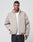 A man stands facing forward in the light beige LIVE FAST DIE YOUNG LFDY Puffer Jacket with a high collar and subtle logo stitching, worn over a white T-shirt and dark jeans, left hand in pocket, against a plain light background.