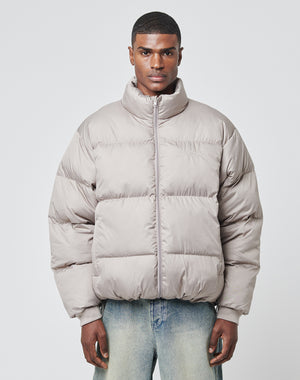 A man faces forward wearing an oversize fit light gray LFDY Puffer Jacket by LIVE FAST DIE YOUNG, featuring logo stitching, paired with faded blue jeans against a plain light background.