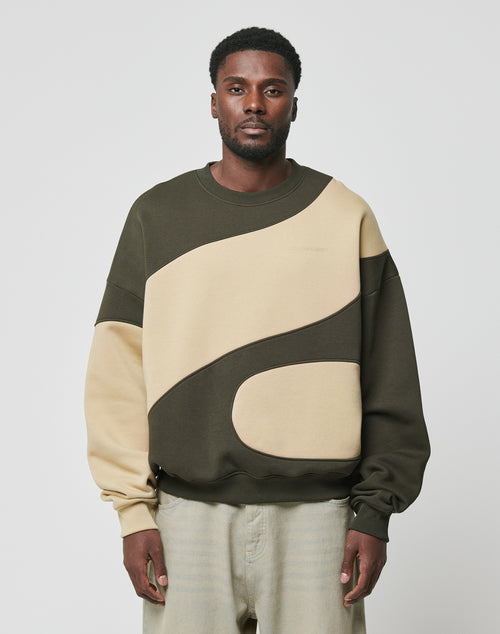 A man faces forward in a LIVE FAST DIE YOUNG LFDY Wavy Sweater, an oversized two-tone olive green and beige Baumwolle piece with curved panel designs, paired with light gray pants against a plain background.