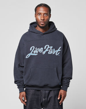 A man stands facing the camera in a dark blue LFDY Script Hooded sweatshirt from LIVE FAST DIE YOUNG, featuring “LivePart” in large script on the front. The oversized fit adds comfort and style against a light gray background.