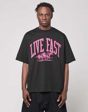 Wearing the Live Fast Stallion Tee by LIVE FAST DIE YOUNG—a black oversize 100% Baumwolle shirt with bold pink “LIVE FAST” frontprint above two horses—the person stands against a plain light gray background, facing forward neutrally.