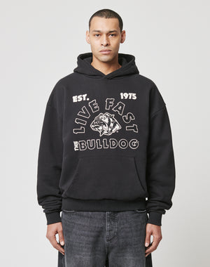 A person in a black LFDY The Bulldog Hooded by LIVE FAST DIE YOUNG, featuring an embroidered EST. 1975 LIVE FAST THE BULLDOG and bulldog graphic, paired with dark jeans, stands against a plain white background.