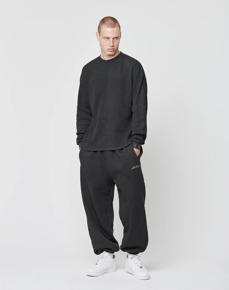 A man with short hair stands against a plain background, wearing the LIVE FAST DIE YOUNG Blank Waffle Longsleeve in black and matching black sweatpants aus 100% Baumwolle. He pairs the look with white sneakers, gazing slightly downward with hands in his pockets.