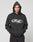 Someone stands against a plain light gray background wearing the LFDY Sphere Hooded by LIVE FAST DIE YOUNG—an oversized black hoodie featuring a silver LIVE FAST. front print, with the hood up.