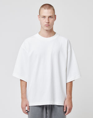 A young man with short blond hair wears a plain oversized white Cropped Oversize Tee by LIVE FAST DIE YOUNG and gray pants, standing against a light gray background. He faces the camera with a neutral expression; the tee features a small Flag-Label accent.
