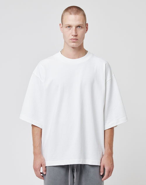 A young man with short blond hair wears a plain oversized white Cropped Oversize Tee by LIVE FAST DIE YOUNG and gray pants, standing against a light gray background. He faces the camera with a neutral expression; the tee features a small Flag-Label accent.