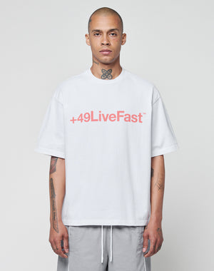 A person with a shaved head faces forward against a plain light background, wearing the LIVE FAST DIE YOUNG Basic +49 Tee—an oversized white cotton shirt featuring a red +49LiveFast front print—paired with gray shorts and showing neck and arm tattoos.