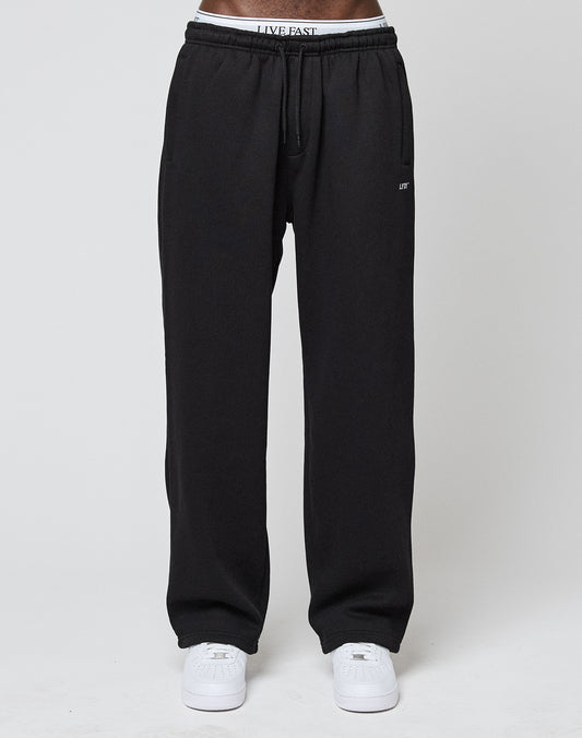 A person wears LIVE FAST DIE YOUNG Basic Wide Jogger sweatpants with an elastic waistband and logo print, partially revealing white underwear with LIVE FAST lettering, plus white sneakers, standing against a plain light background.