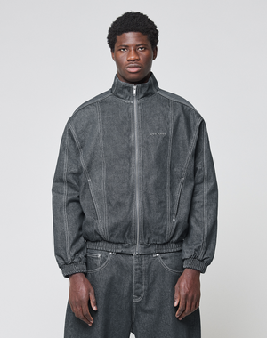 A man faces forward in a dark gray LFDY Denim Trackjacket by LIVE FAST DIE YOUNG, with visible seams, subtle chest text, and paired baggy pants. The 100% cotton jacket features a stand-up collar and is set against a plain light background.