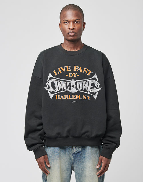 A person wears the LIVE FAST DIE YOUNG Live Fast Tribal Sweater, an oversize fit black cotton sweatshirt with a striking tribal graphic and LIVE FAST DIY HARLEM, NY on the front, paired with light blue jeans against a plain white background.