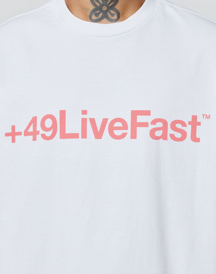 Someone wears the LIVE FAST DIE YOUNG Basic +49 Tee—an oversized white shirt with a bold pink +49LiveFast™ front print, crafted from soft Baumwolle for comfort. A floral or abstract neck tattoo shows above the collar.