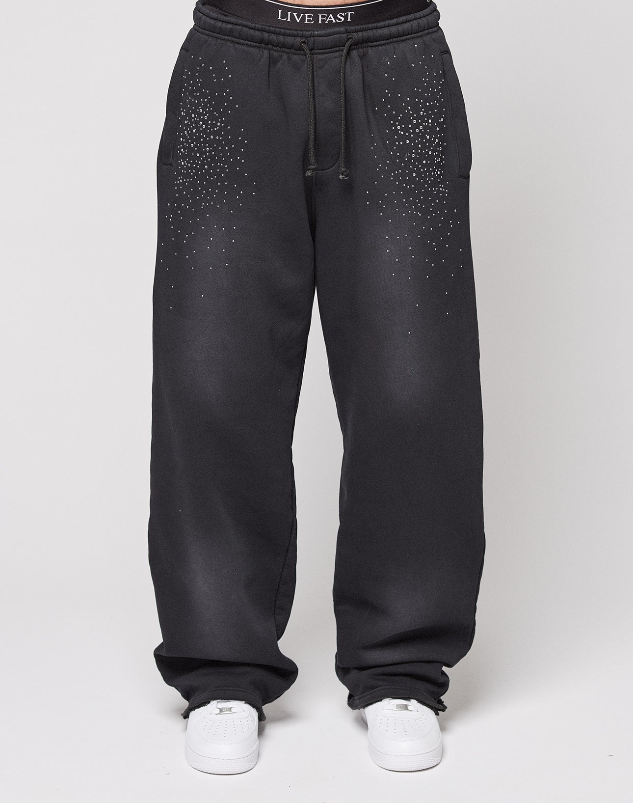A person wearing LIVE FAST DIE YOUNG's Live Fast Rhinestone Jogger—wide fit black sweatpants with white dotted pocket details, rhinestones, white sneakers, and a visible “LIVE FAST” waistband.