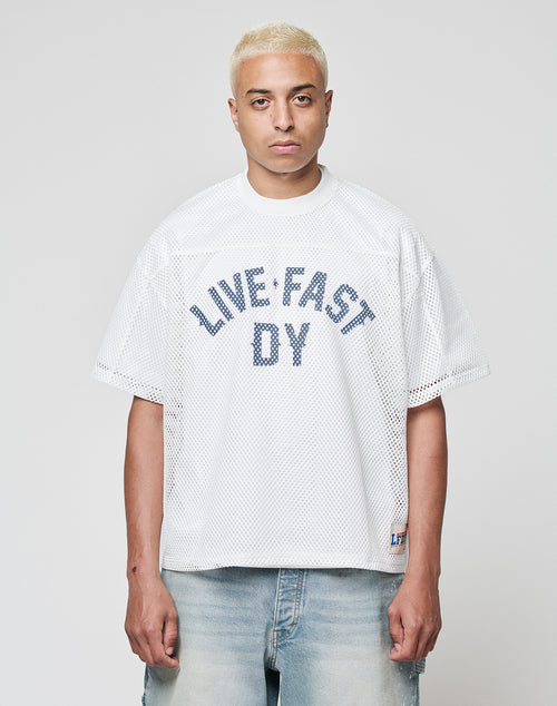 A person with short, bleached blond hair models the LIVE FAST DIE YOUNG LFDY Boxy Jersey—a loose white mesh shirt with a bold blue LIVE FAST DY front print—paired with light denim jeans, standing before a plain light gray backdrop.