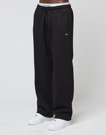 A person stands against a plain white background, wearing LIVE FAST DIE YOUNGs Basic Wide Jogger in black (loose fit), white sneakers, and a visible white waistband with the LIVE FAST logo.