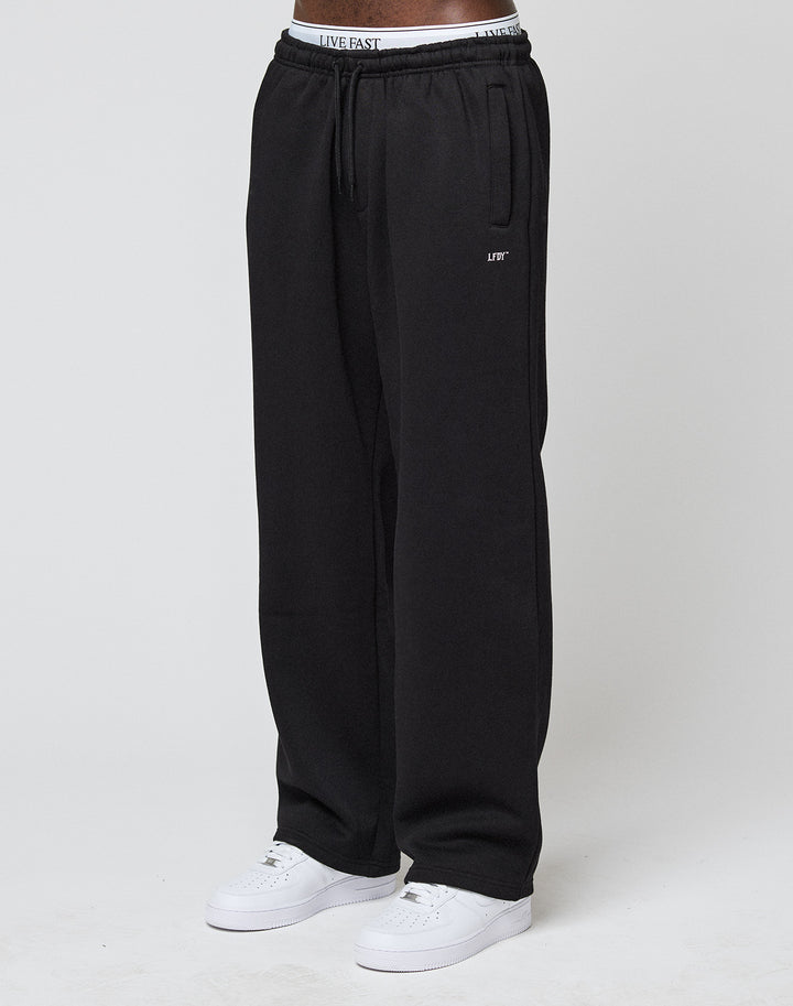 A person stands against a plain white background, wearing LIVE FAST DIE YOUNGs Basic Wide Jogger in black (loose fit), white sneakers, and a visible white waistband with the LIVE FAST logo.