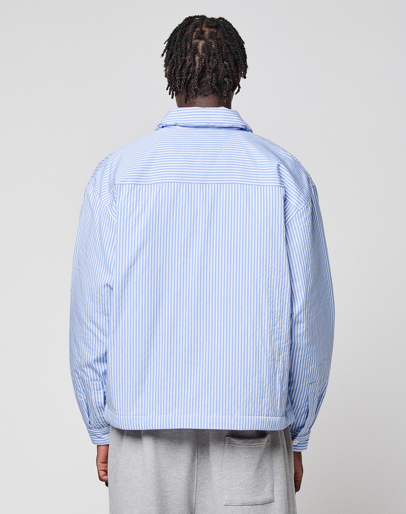 A person with short braids, seen from behind, wears the LFDY Striped Padded Shirt by LIVE FAST DIE YOUNG in light blue and white vertical stripes with brusttaschen and light gray pants against a plain, light background.