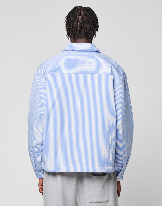 A person with short braids, seen from behind, wears the LFDY Striped Padded Shirt by LIVE FAST DIE YOUNG in light blue and white vertical stripes with brusttaschen and light gray pants against a plain, light background.