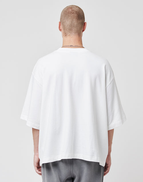 Seen from behind, a person with short hair wears the LIVE FAST DIE YOUNG Cropped Oversize Tee in soft Baumwolle and dark gray pants. The subtle flag-label adds a modern touch against the plain light background.