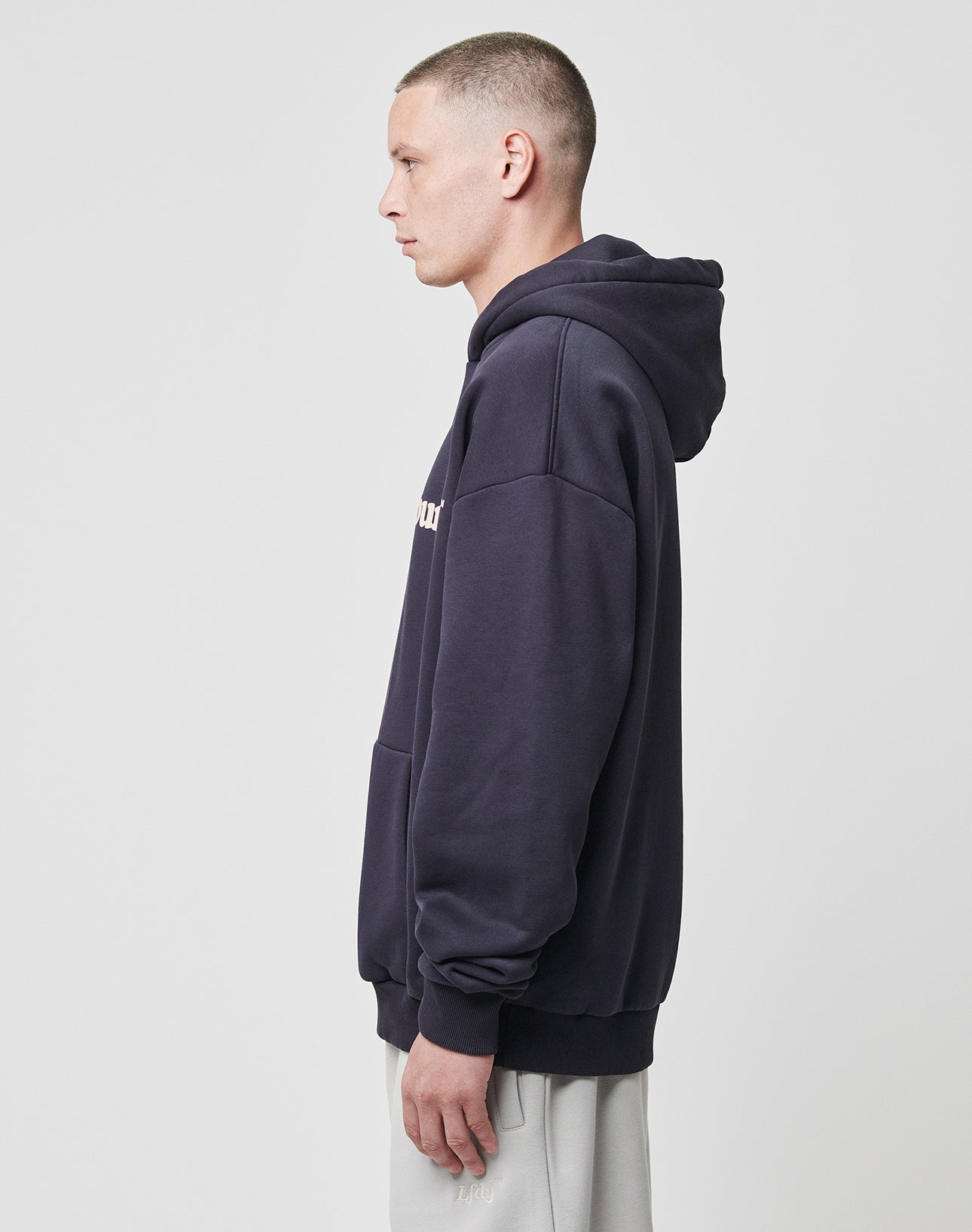 A person with short hair wears the LIVE FAST DIE YOUNG Basic Chest Hooded in an oversized fit and light pants, standing in profile against a plain white background.