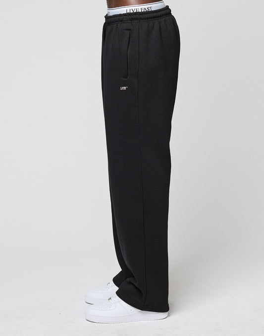 A person stands sideways against a plain light background wearing LIVE FAST DIE YOUNG Basic Wide Jogger black sweatpants with LIFT logo on the thigh, a white LIVE FAST elastic waistband, and white sneakers.