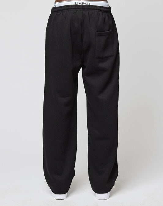 A person is shown from the back wearing LIVE FAST DIE YOUNG Basic Wide Jogger sweatpants in black with an oversize fit, elastic waistband, back pocket, white visible waistband, and white shoes against a plain light gray background.