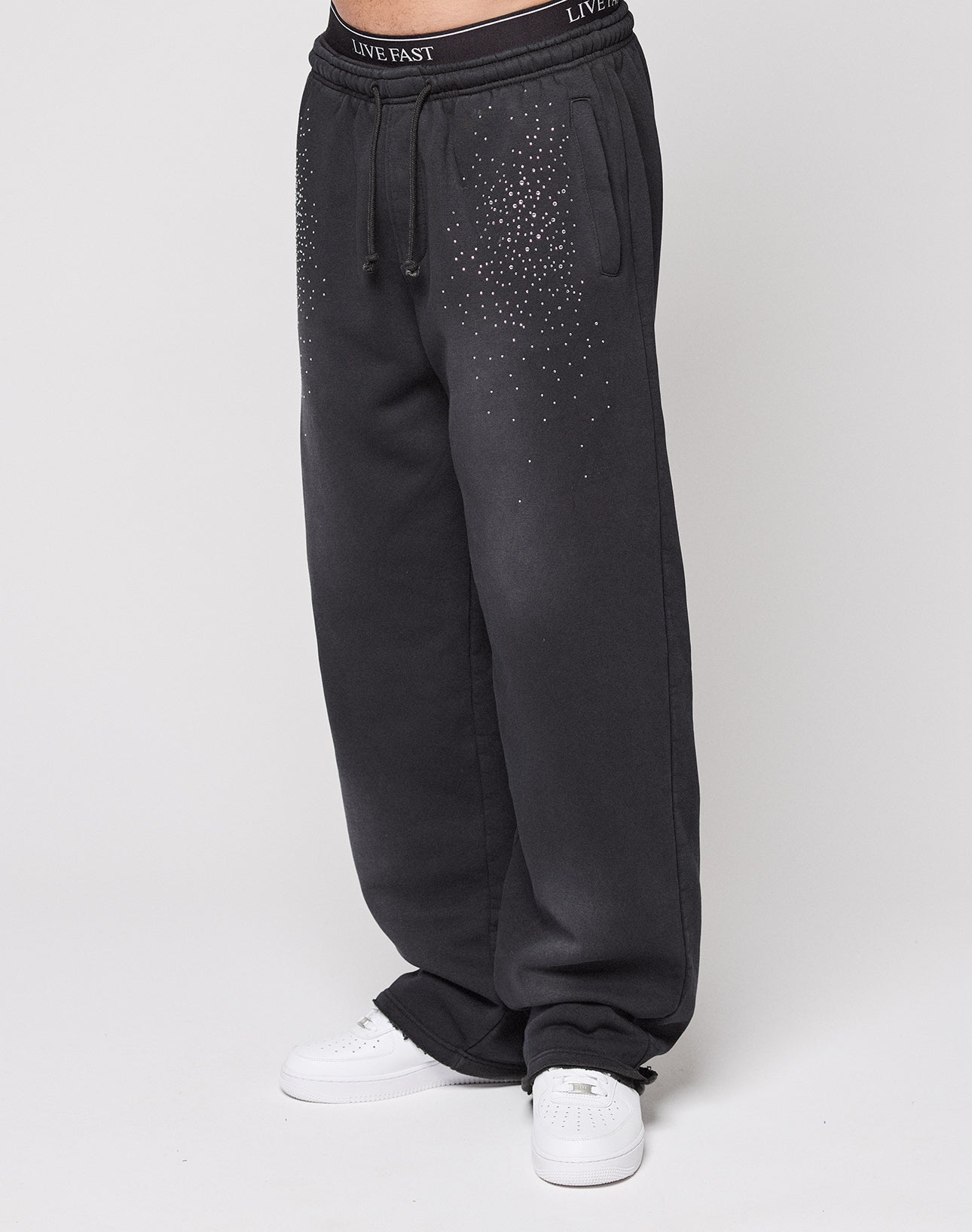 Someone stands against a plain light background wearing the LIVE FAST DIE YOUNG Live Fast Rhinestone Jogger—wide fit black sweatpants with small white rhinestone embellishments on the hips and a black LIVE FAST waistband—paired with white sneakers.