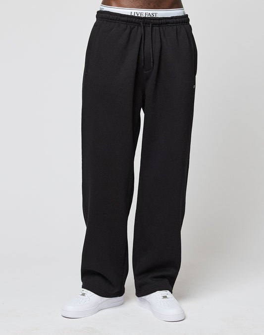 A person wears Basic Wide Jogger sweatpants by LIVE FAST DIE YOUNG, paired with white sneakers and logo waistband underwear reading LIVE FAST, standing against a plain white background.
