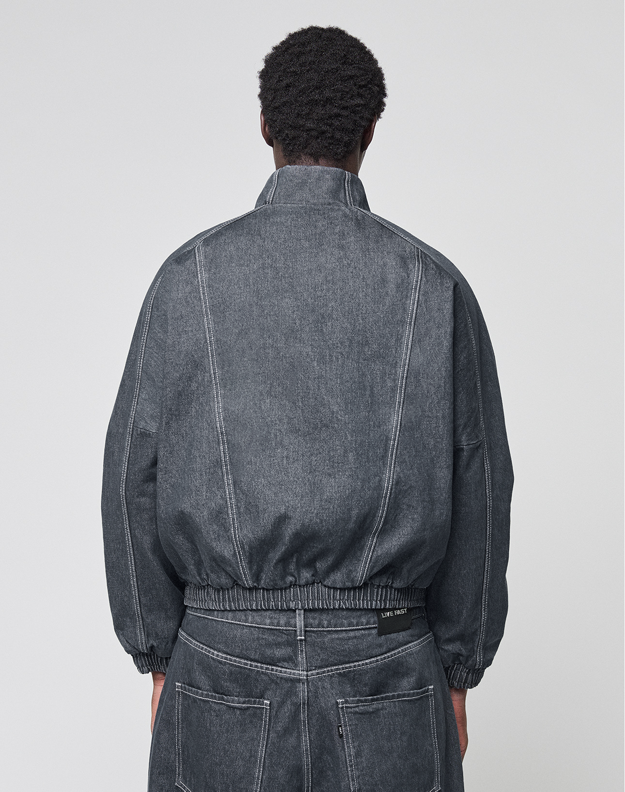 Seen from behind, a person with short curly hair wears the LIVE FAST DIE YOUNG LFDY Denim Trackjacket, featuring a Stehkragen and elastic waistband, made from 100% cotton, paired with matching denim pants against a plain light background.