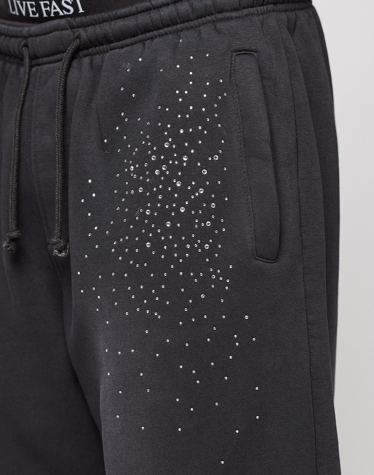 The Live Fast Rhinestone Jogger by LIVE FAST DIE YOUNG features black sweatpants with an elastic waistband, side pocket, rhinestone Muster on the upper thigh, partially visible "LIVE FAST" text, and a wide fit for comfort.
