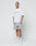 A person with tattoos stands against a plain white background wearing the LIVE FAST DIE YOUNG Basic +49 Tee in white with red frontprint, gray shorts, white crew socks, and white sneakers, showing a neutral expression.