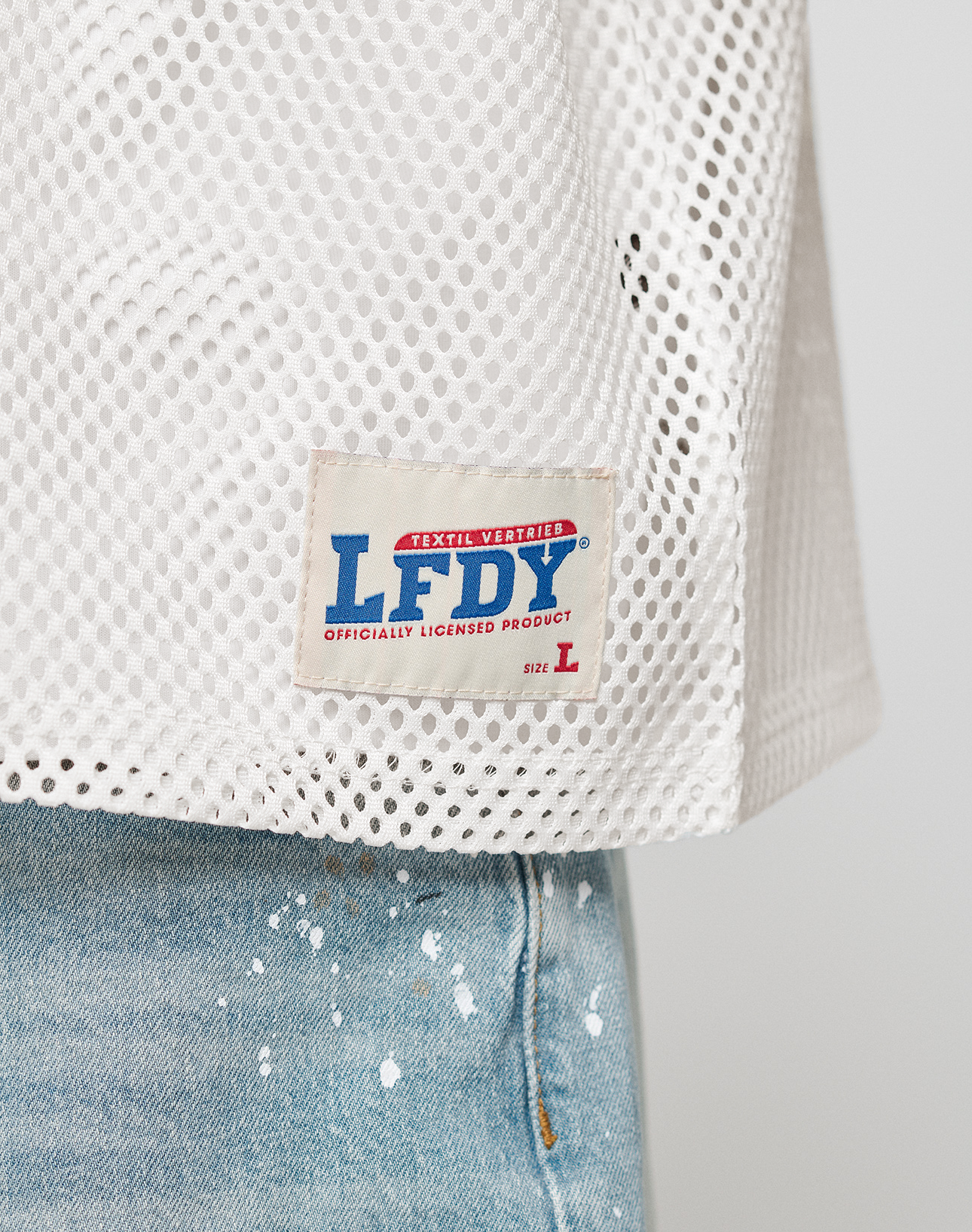Close-up of a white LFDY Boxy Jersey by LIVE FAST DIE YOUNG with a label near the hem, styled with light blue jeans featuring white paint splatters.